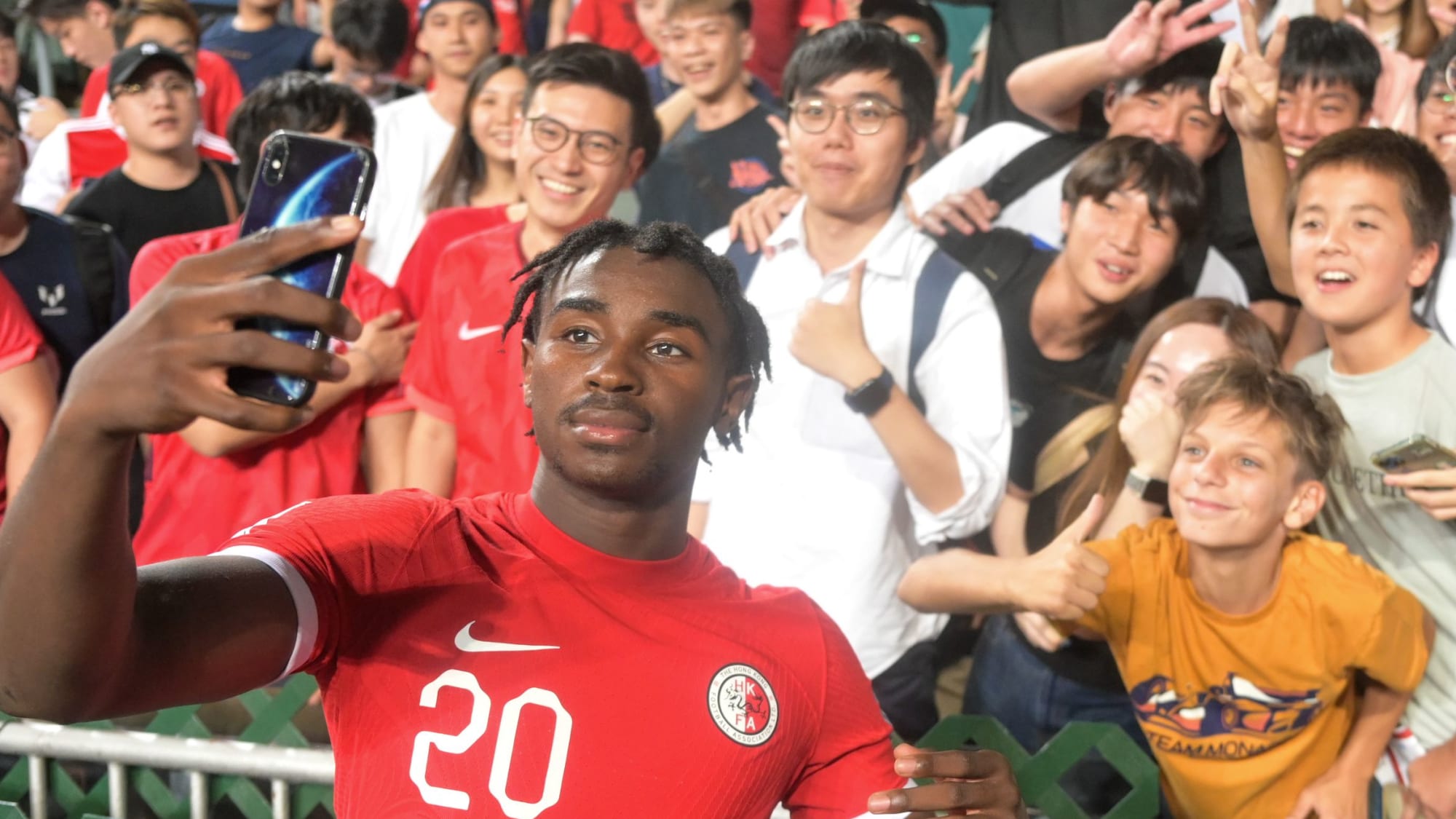 Michael Udebuluzor feiert im Trikot von Hongkongs Nationalmannschaft mit den Fans und macht ein Handy-Selfie.