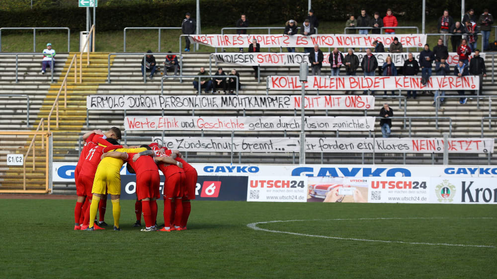 Unruhige Zeiten: Vorne stimmt sich die Mannschaft des 1. FC Kaan-Marienborn auf das Derby ein, im Hintergrund protestieren Fans der Sportfreunde Siegen gegen die Fusionspl&#228;ne.